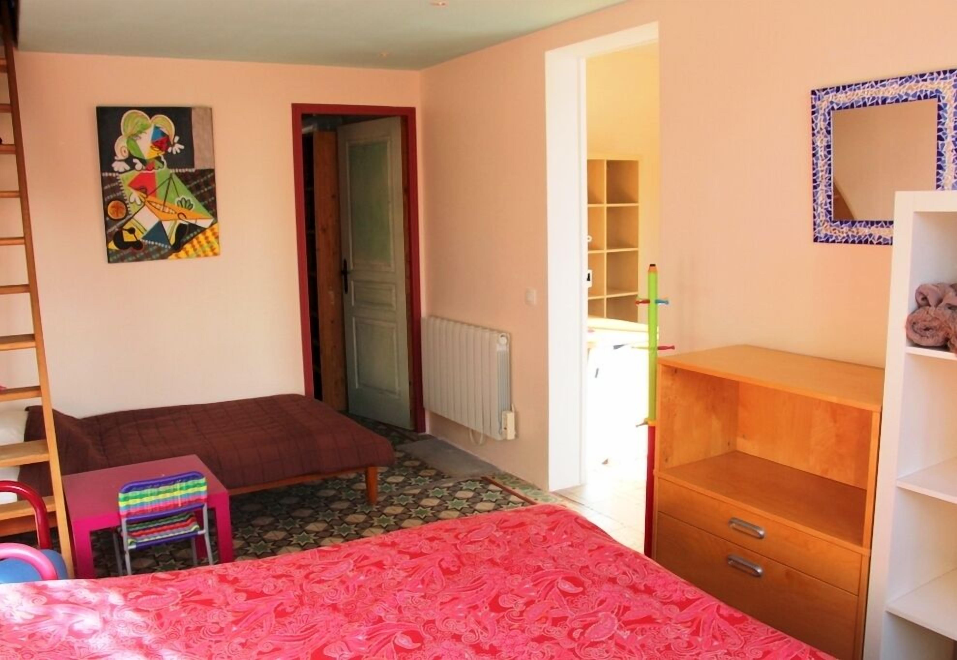 Une chambre à coucher comprenant un lit, une commode en bois, une chaise, un radiateur et une porte donnant sur une autre pièce.