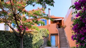 Ein schmaler Gang mit einem Kiesweg, ein Baum mit rosa Blüten und ein Gebäude mit einer blauen Tür sowie einer Treppe zu einem überdachten Bereich.