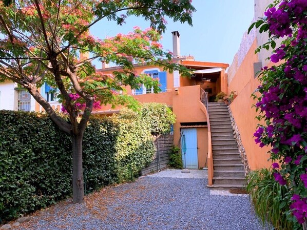 A narrow alleyway with a gravel path, a tree with pink flowers, and a building with a blue door and a staircase leading to a covered area.
