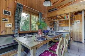 A wooden interior with a dining table set for four, a kitchen area, and a window with a view of trees. - Chalet l'Ange - La Tzoumaz, 4 Vallees (La Tzoumaz)