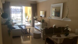 A living room with a sofa, coffee table, dining table, and chairs. There is a window with curtains, a lamp, and a framed picture on the wall. - Estepona, cozy 2 bedrooms apartment close to the beach and marina (Estepona)