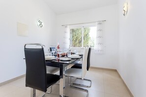 A dining area with a table set for two, chairs, a window with curtains, and a wall-mounted light fixture. - CoolHouses Algarve Lagos | Casa Redonda (Lagos)