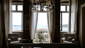 A room with a view of the sea, featuring two armchairs, a sofa, a chandelier, and a table with books.