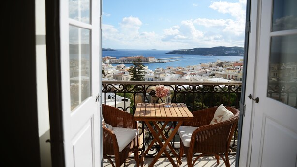 An open balcony with wicker furniture and a table, overlooking a coastal town with buildings and a pier.