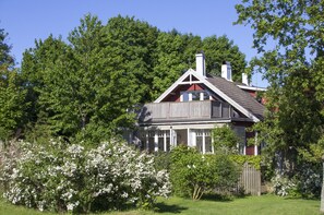 Et træhus med saddeltag, omgivet af frodig vegetation og blomstrende buske.