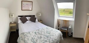 A neatly made bed with a floral duvet, two bedside lamps, a framed picture on the wall, and a view of the outdoors through a window. - Traditional Croft House with garden. Ample parking and central location. (Isle of Skye)