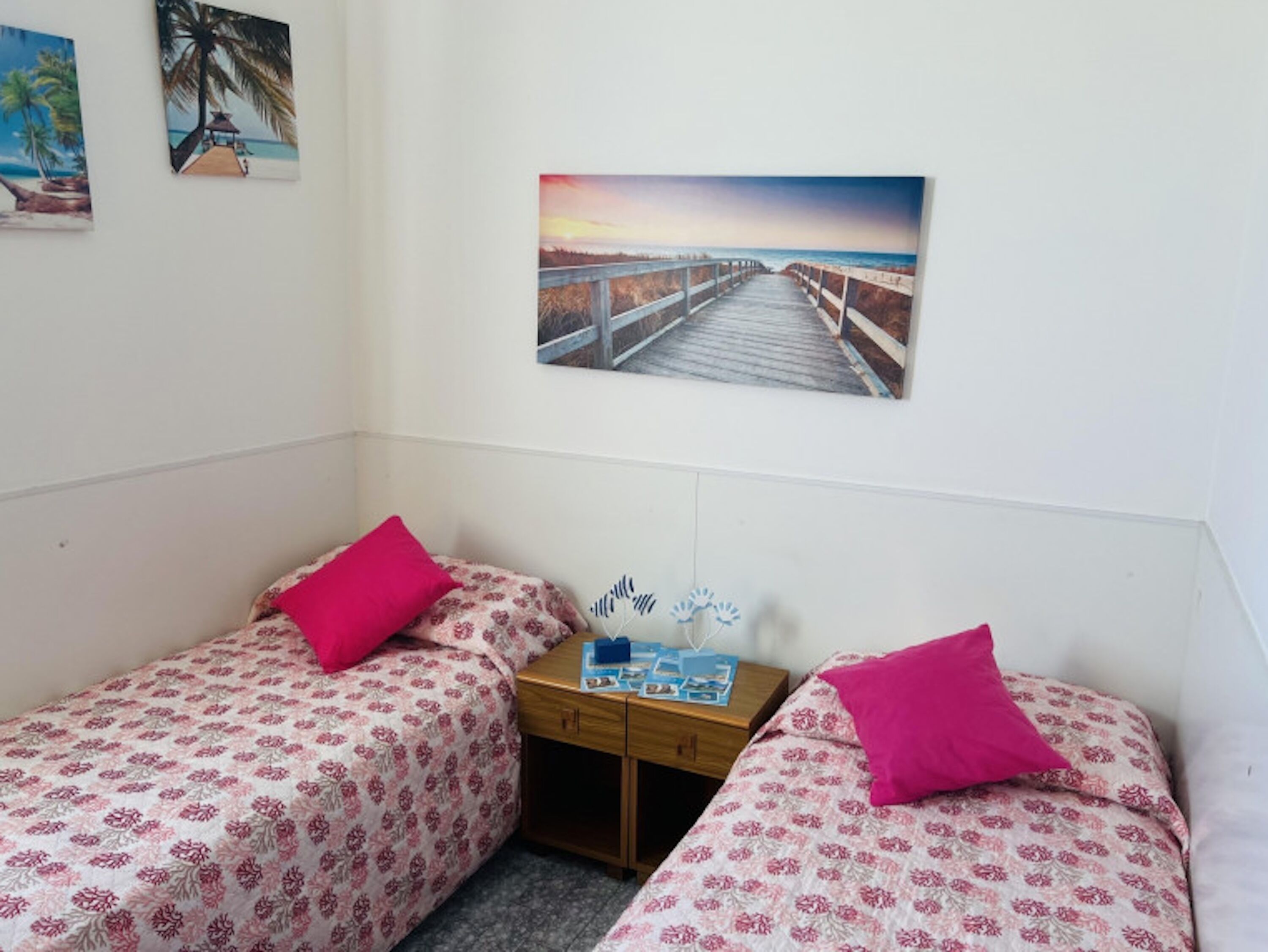 A room with two single beds, each with a pink pillow and a floral-patterned bedspread. There is a wooden nightstand with a small table between the beds, and a framed picture of a pier on the wall above the nightstand.