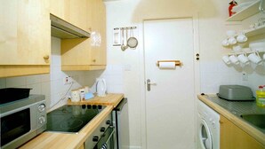A compact kitchen with a microwave, stove, oven, and washing machine. There is a door with a towel holder and a shelf with cups.