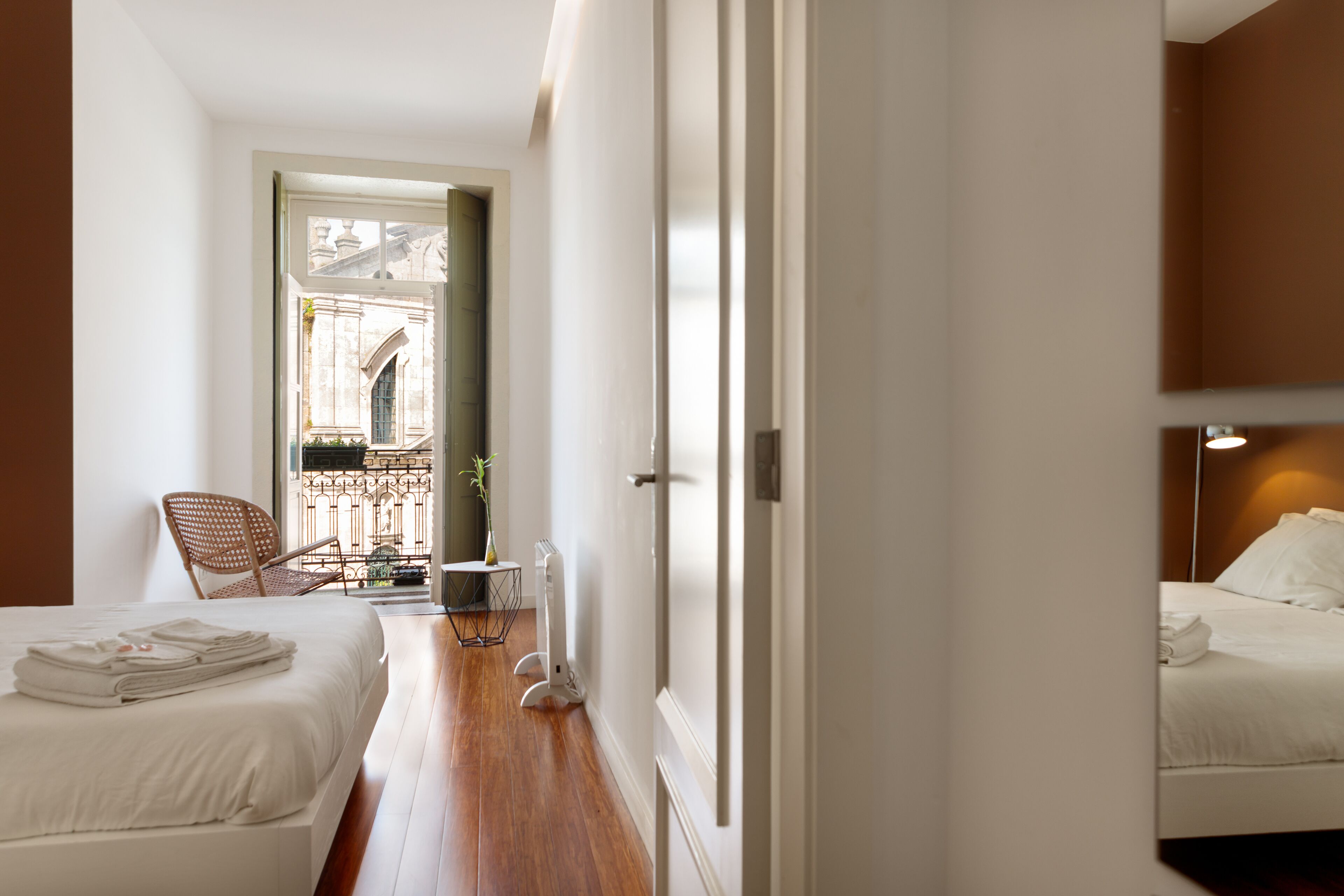 A hotel room with a bed, a chair, a small table with a vase, and a view of a building through an open door.