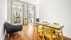Una sala moderna con un sofá, una mesa de comedor y sillas, ventanas amplias con vista a un edificio, y un taburete con una planta.