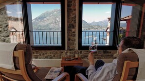 Un homme se détend sur une chaise en bois près d’une fenêtre, d’où l’on peut admirer un paysage de montagne et de lac.