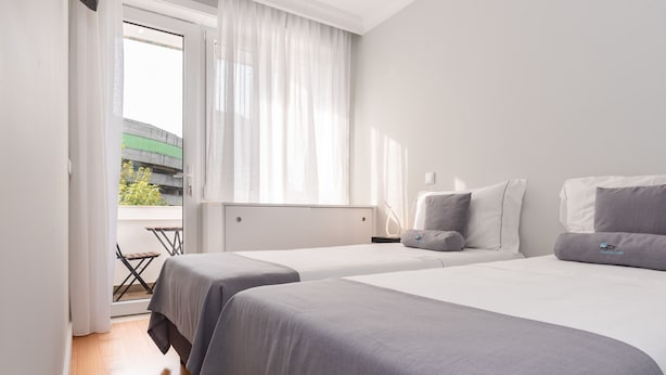 Ein Hotelzimmer mit zwei Betten, einem Fenster mit Vorhängen, Blick auf ein Gebäude und einem kleinen Tisch am Fenster.