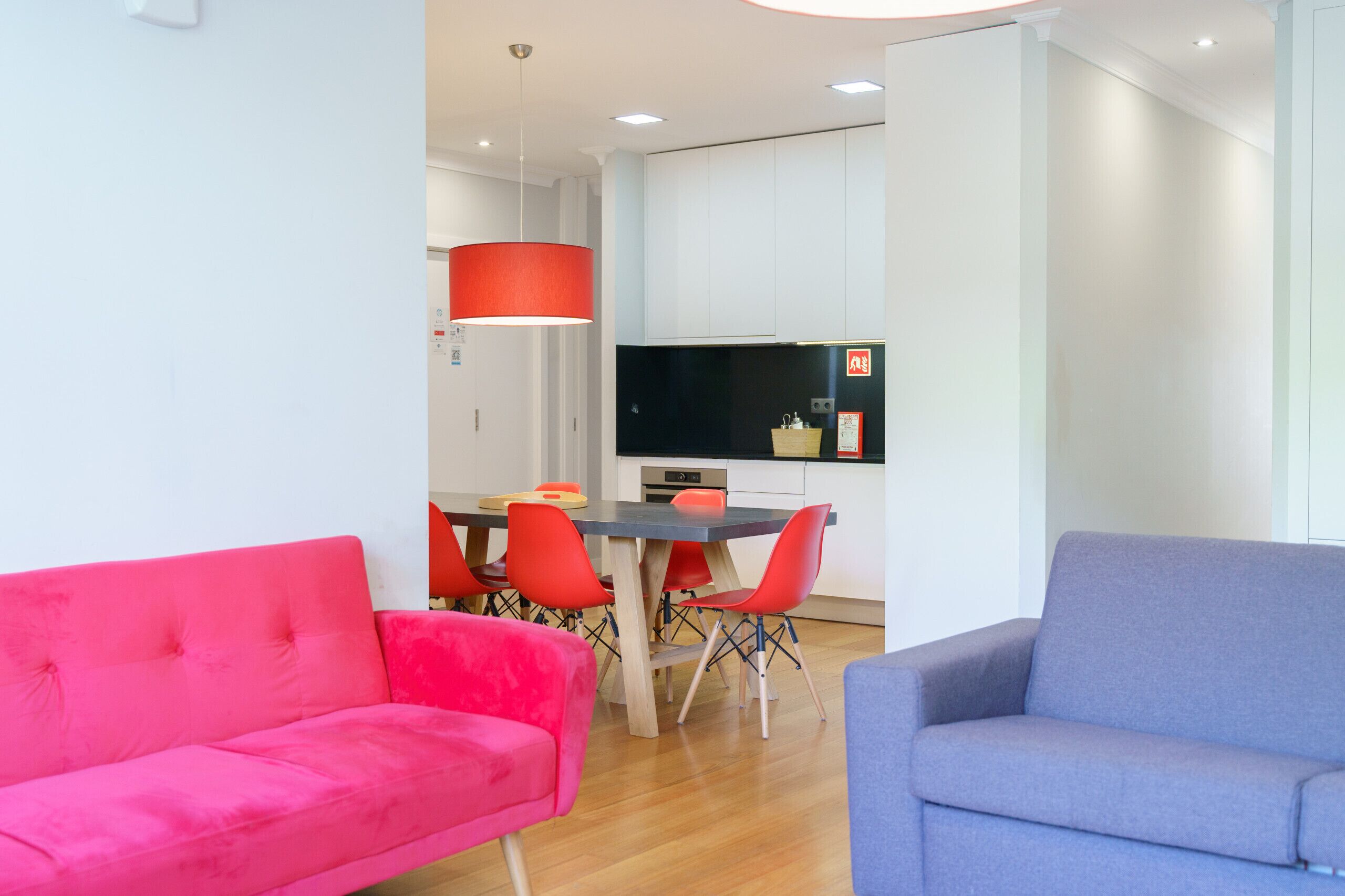 Uma sala de estar moderna com um sofá vermelho vibrante, um sofá azul e uma área de jantar com cadeiras vermelhas e uma mesa preta.