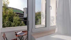 Ein Balkon mit Tisch und Stühlen, ein Fenster mit Vorhängen und Blick auf ein Gebäude und Bäume.