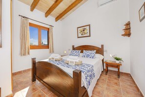 A bedroom with a wooden bed, a bedside table with a lamp, a window with curtains, and a wall-mounted shelf.