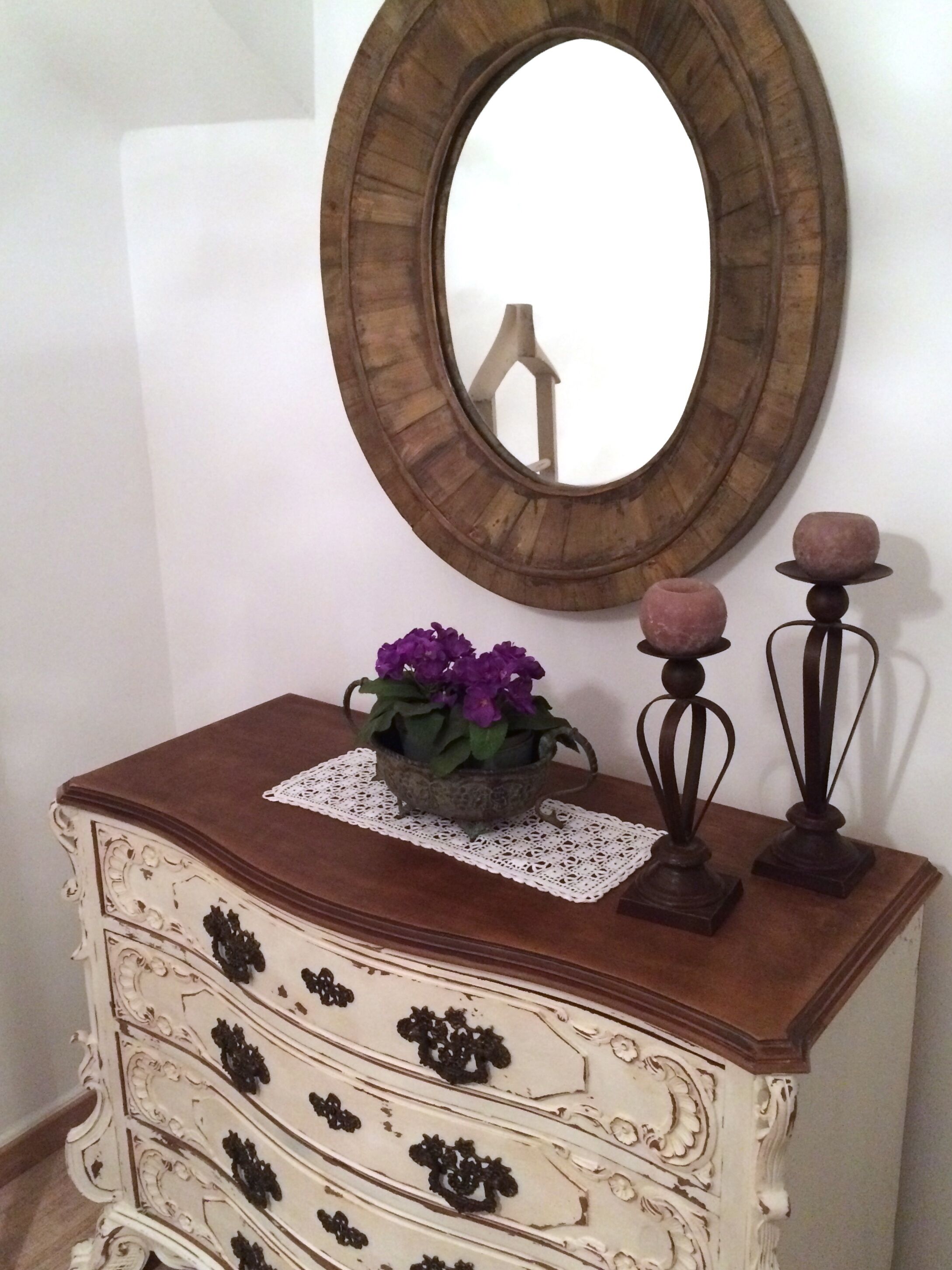 Eine Holzkommode mit einem Spiegel darüber, einer Spitzendecke und einem Blumentopf mit violetten Blüten.