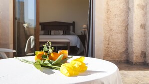 Una mesa con un ramo de flores amarillas y un vaso de jugo de naranja, en un entorno exterior con vista a una cama y una ventana.