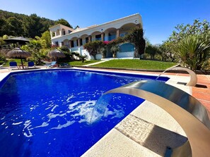 Una piscina con superficie de agua azul cristalina, rodeada por una zona de patio con sillones de descanso y una estructura de techo de paja.