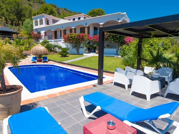 Una piscina con sillones de descanso, una pérgola y una casa al fondo.