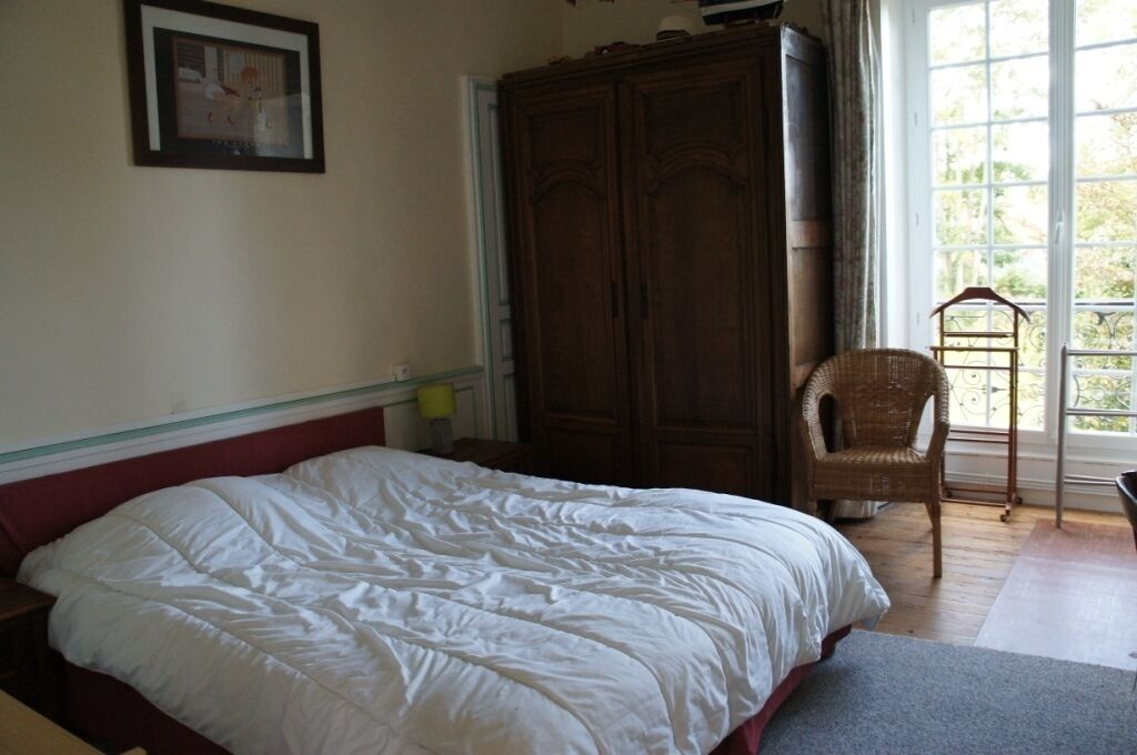 Een slaapkamer met een groot bed, een houten garderobe, een rieten stoel en een raam met uitzicht op groen.