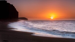 Uma praia com pôr do sol, ondas e uma formação rochosa.