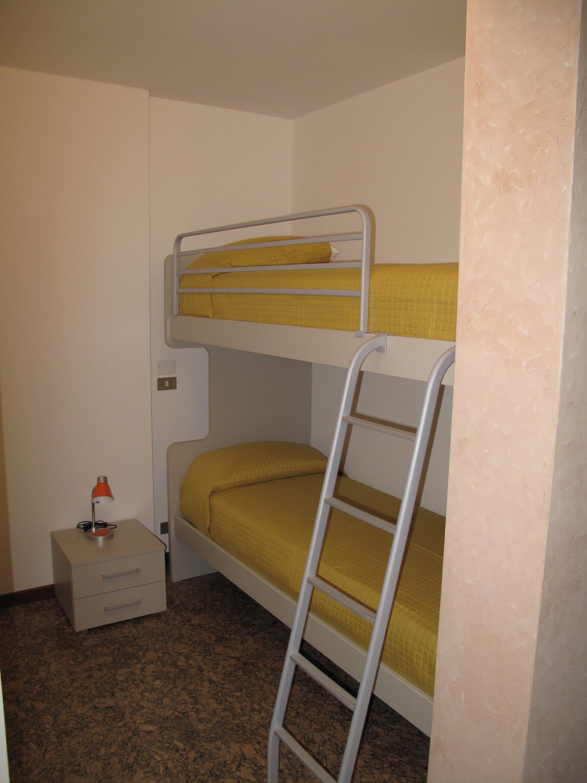 A bunk bed room with a ladder, a small drawer unit, and a desk with a lamp.