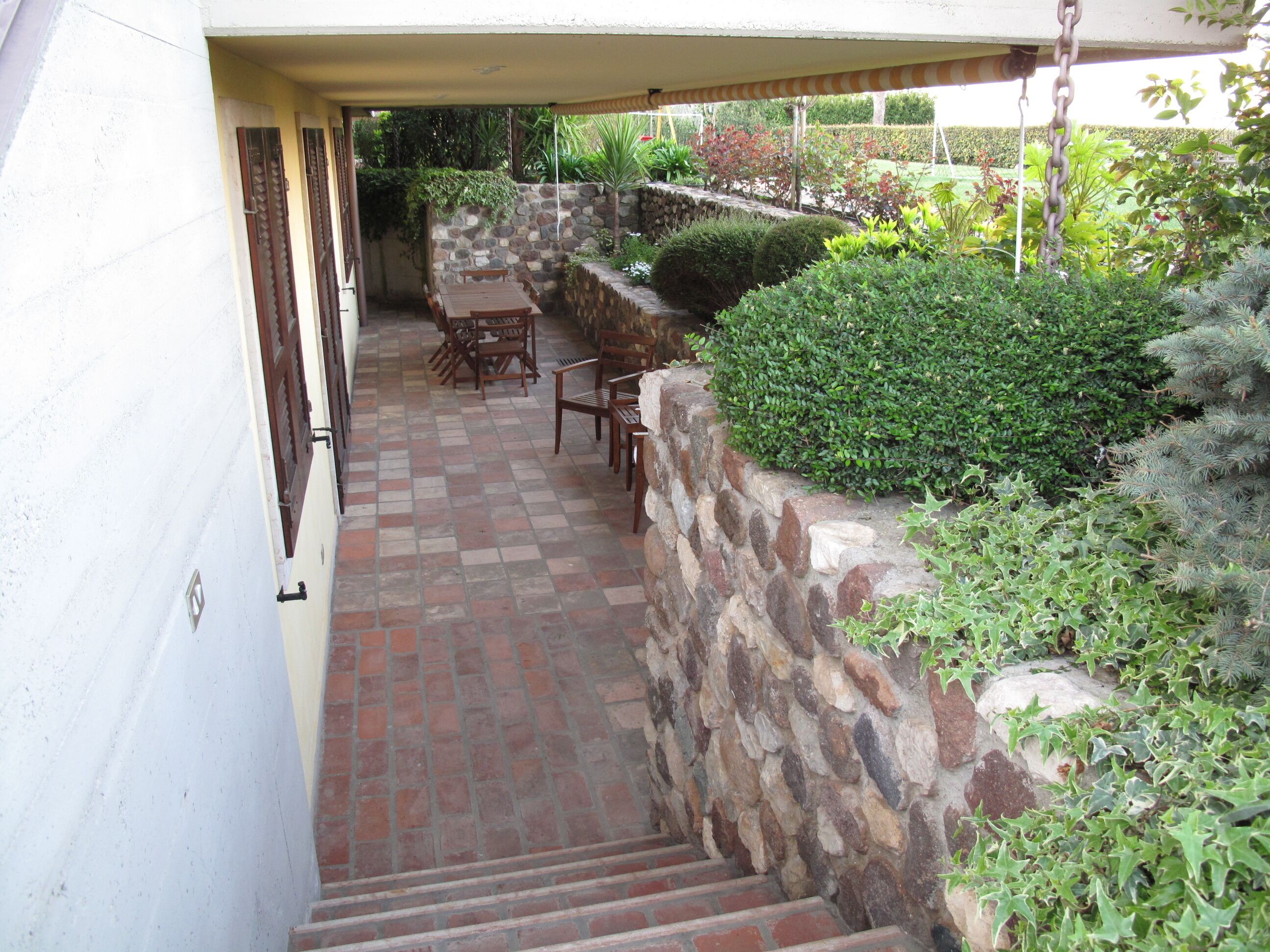 A covered patio with a stone wall, outdoor furniture, and a staircase leading to a higher level.