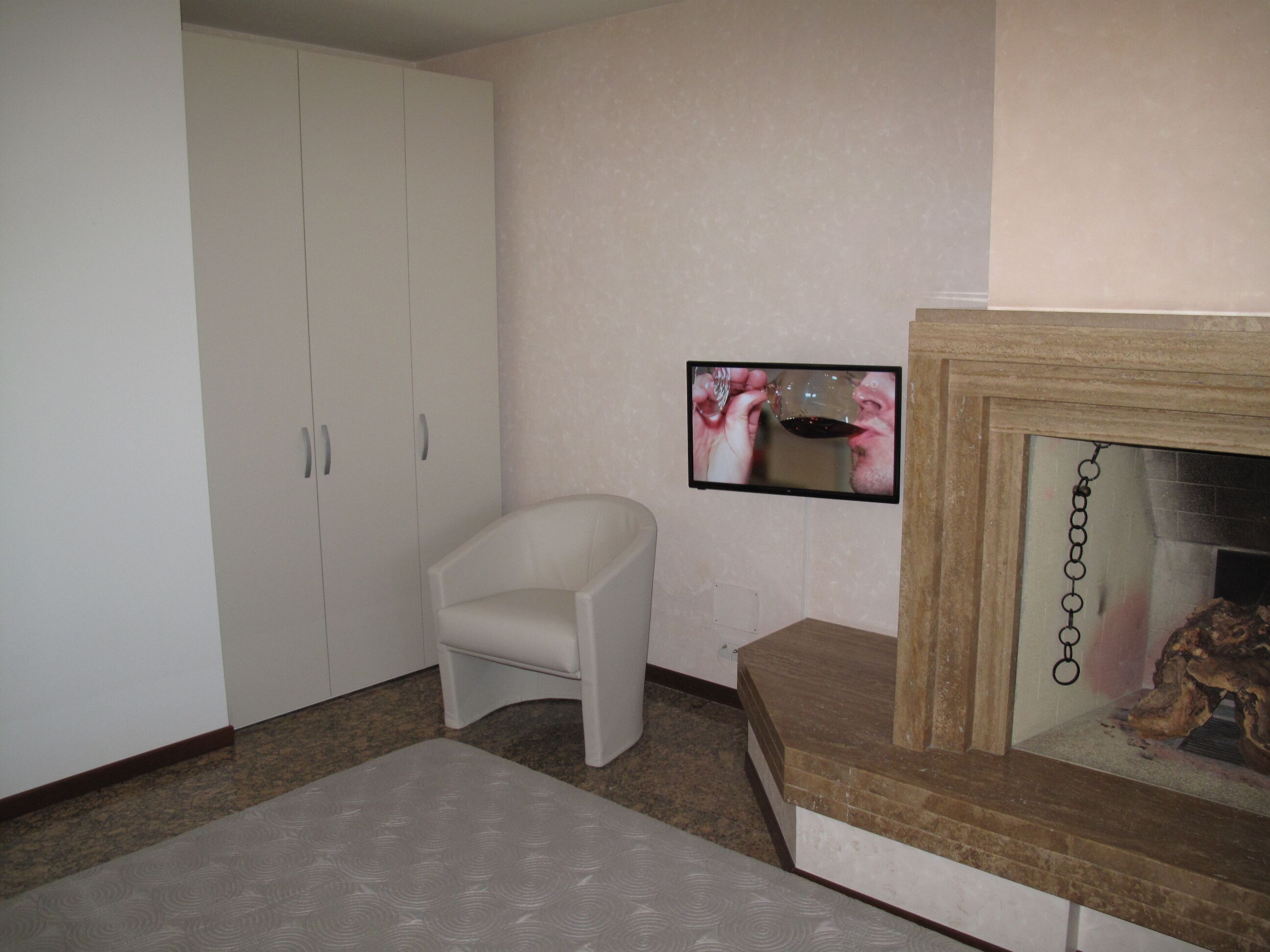 Une pièce avec une armoire blanche, un petit fauteuil blanc, un téléviseur à écran plat fixé au mur et une cheminée avec un manteau en pierre.