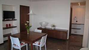 A modern dining area with a wooden table, white chairs, a TV on a shelf, and a pendant light. - APARTMENT INSIDE A VILLA, LARGE GARDEN WITH LAKE AND POOL VIEW (Bardolino)