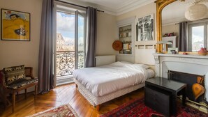 A bedroom with a bed, a dresser, a chair, a fireplace, and a view of a building through a window.