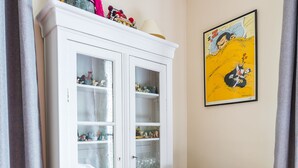 A room with a white cabinet with glass doors, a patterned rug, a wooden chair with an elephant design, and a yellow framed picture on the wall.