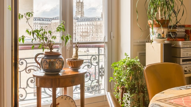 A room with a large window, a wooden table with decorative items, a chair, and a view of a cityscape.