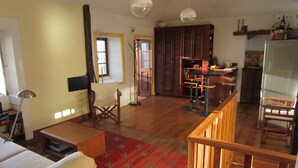 A spacious living area with a wooden floor, a red patterned rug, a white sofa, a wooden coffee table, and a wooden chair. There is a kitchen area with a refrigerator, a sink, and a countertop. - Villa with private pool, large garden and stunning sea view (Faja da Ovelha)