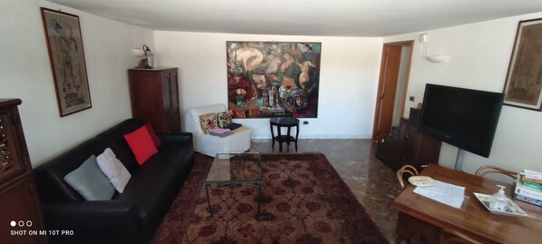Een woonkamer met een zwarte bank, een glazen salontafel, een rood kussen, een groot abstract schilderij, een flatscreen televisie en een houten bijzettafeltje met een dienblad.