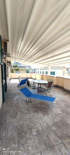 Een overdekt terras met een tafel, stoelen, een blauwe zonnescherm en een betegelde vloer.