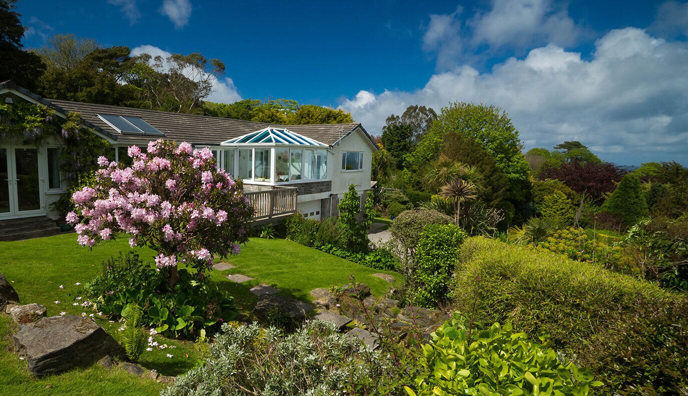 Een huis met een tuinkamer, omgeven door een verzorgde tuin met diverse planten en een grote bloeiende roze boom.