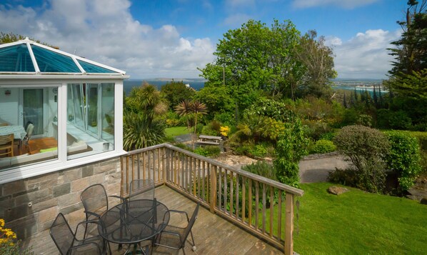 Un espace extérieur abrité, doté d’une table et de chaises, d’un mur en pierre et offrant une vue sur un jardin luxuriant et la côte.