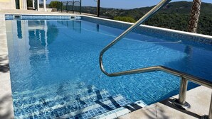 Ein klarer, blauer Infinity-Pool mit einem Metallgeländer, umgeben von einer gefliesten Terrasse und einem Gebäude mit einem Balkon.