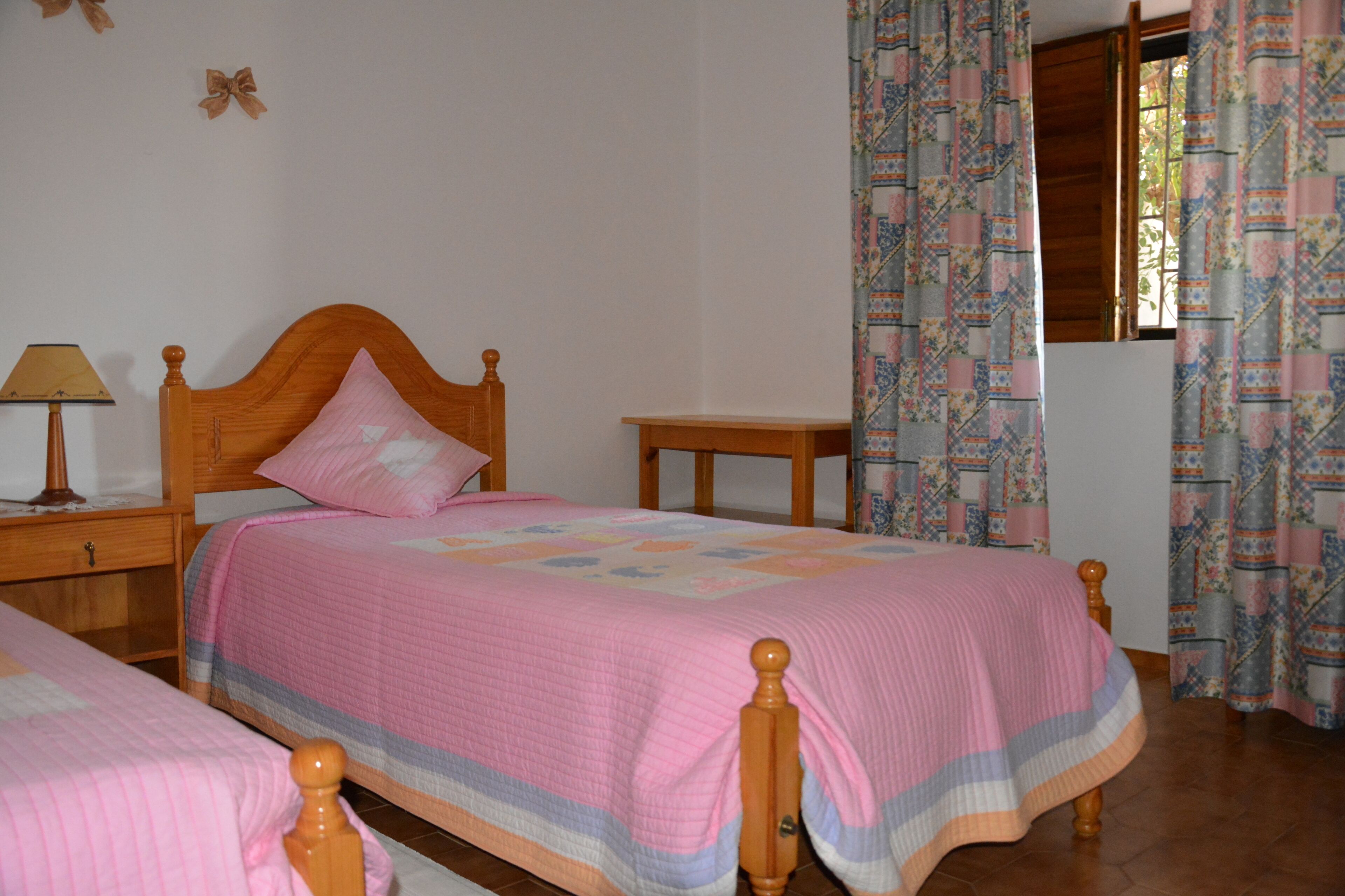 Un dormitorio con dos camas de madera, una mesita de noche con lámpara y una ventana con cortinas estampadas.