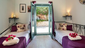 A room with two beds, each with a purple bedspread and floral pillows, a small table with a lamp, and a view of a stone wall and greenery outside. - Artistsvillas-Red Rose,unique,with breathless sea views and ecological pool. (Gaios)