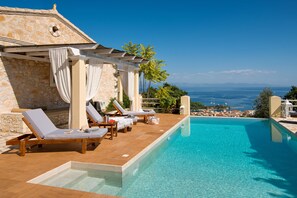 Ein Poolbereich mit Liegestühlen, ein Steinhaus und Blick aufs Meer.