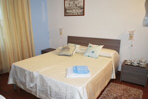 A neatly made bed with a patterned bedspread, pillows with decorative designs, and a bedside table with a lamp and a small tray of tea items.