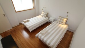 A room with two single beds, wooden flooring, a small white nightstand, and a window with natural light. - OPO-APT 112-D Art Deco apartment in city center. AC, wi-fi, cozy and confortable (Portugal)