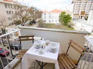 Un balcon avec une table dressée pour deux personnes, offrant une vue sur le paysage urbain avec ses bâtiments et ses arbres.