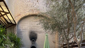 Eine Terrasse mit Holztisch und weißen Stühlen, einem grünen Sonnenschirm und einem Backofen aus Ziegeln.