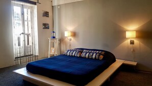 Ein Schlafzimmer mit einem Bett, Nachttischlampen, einem Beistelltisch, einem Fenster mit Vorhängen und einem an der Wand hängenden Bild.