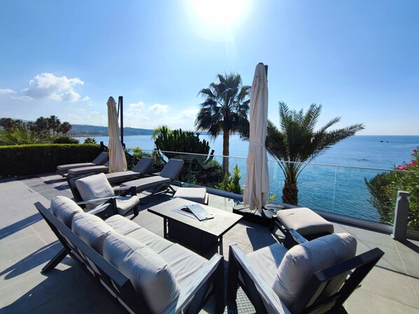A sunlit outdoor seating area with lounge chairs and umbrellas overlooking a body of water.