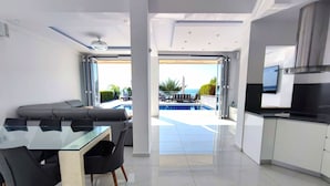 Una sala de estar moderna con una mesa de comedor de vidrio, sillas negras y un sofá gris. Se observa una zona de cocina con encimera negra y armarios blancos. Se ve una piscina a través del espacio abierto.