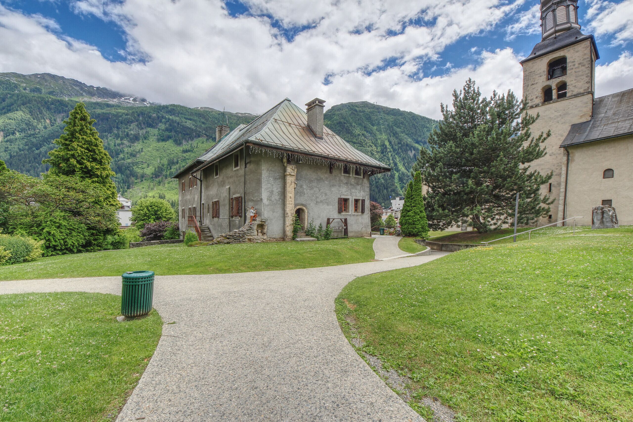 A historic stone building with a steep roof and a bell tower, surrounded by lush greenery and mountains.