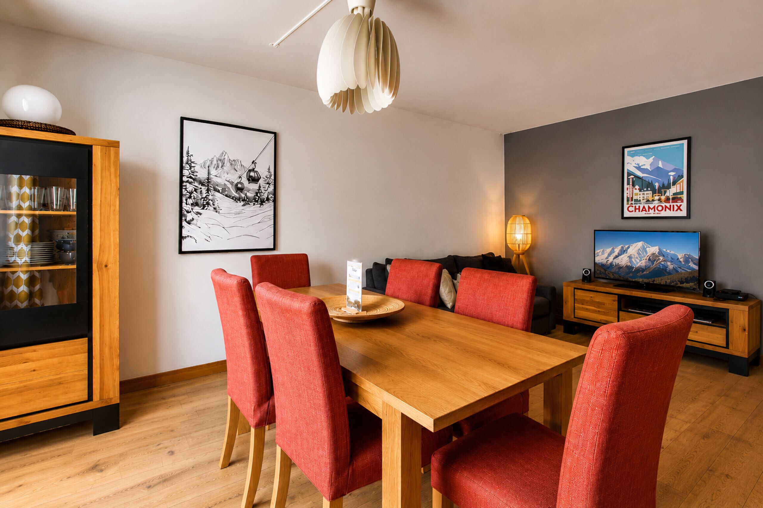 A dining area with a wooden table and red chairs, a cabinet with glassware, and a television on a wooden stand.